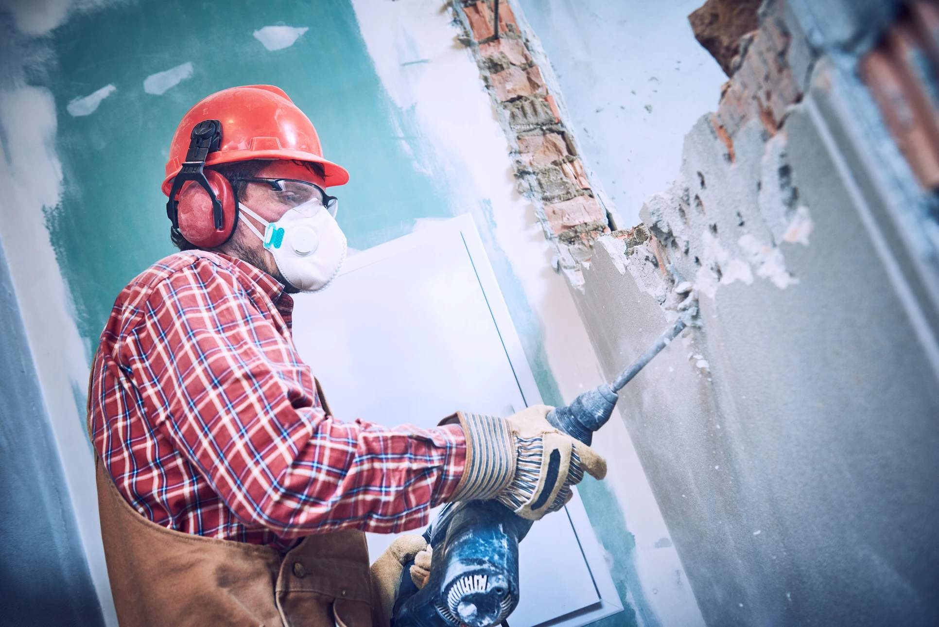Chantier de rénovation intérieur, ouvrier équipé d'un casque rouge, lunettes de protection, masque respiratoire et casque antibruit utilisant un marteau-piqueur pneumatique pour démolir un mur en briques